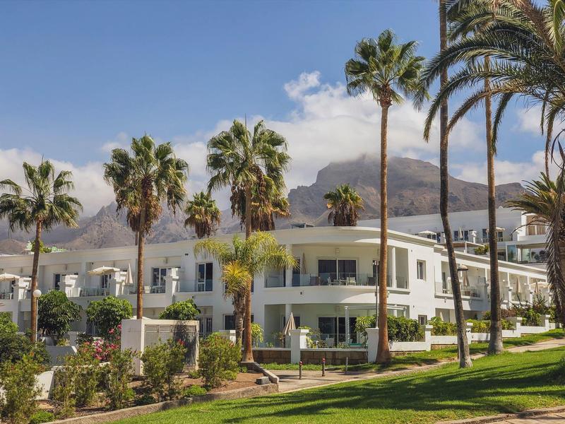 Moderne weiße Gebäude mit großen Fenstern, umgeben von grünen Palmen und Bergen im Hintergrund.