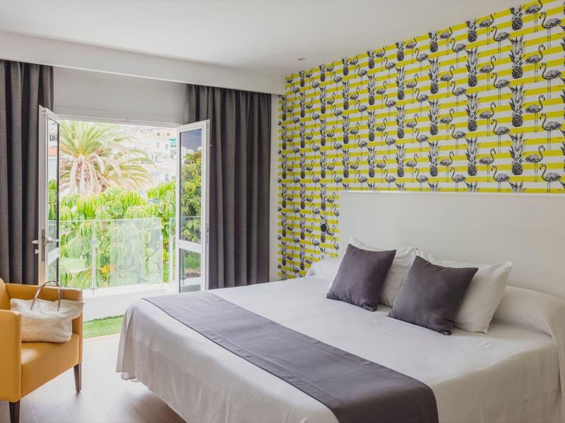 Helles Schlafzimmer mit großem Bett, gelb-gemusterter Wand, grauen Vorhängen und orangem Sessel.