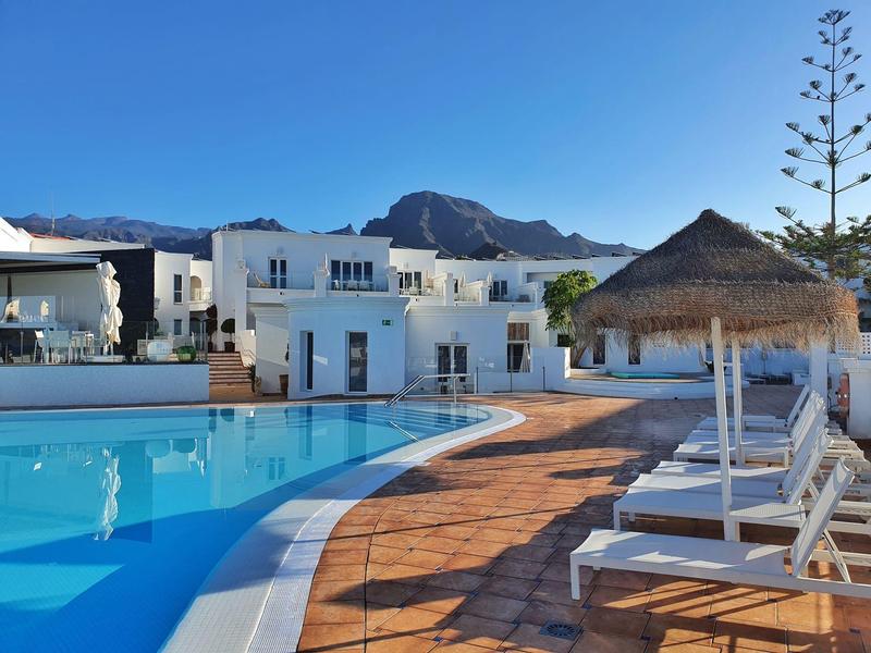 Hotel mit klarem Pool, Liegestühlen und Schirmen vor weißer Gebäudezeile unter blauem Himmel.