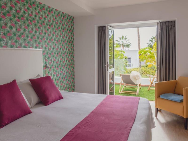 Helles Schlafzimmer mit großem Bett, rosa Akzenten, gemusteter Wand und Blick auf Terrasse mit Gartenmöbeln.