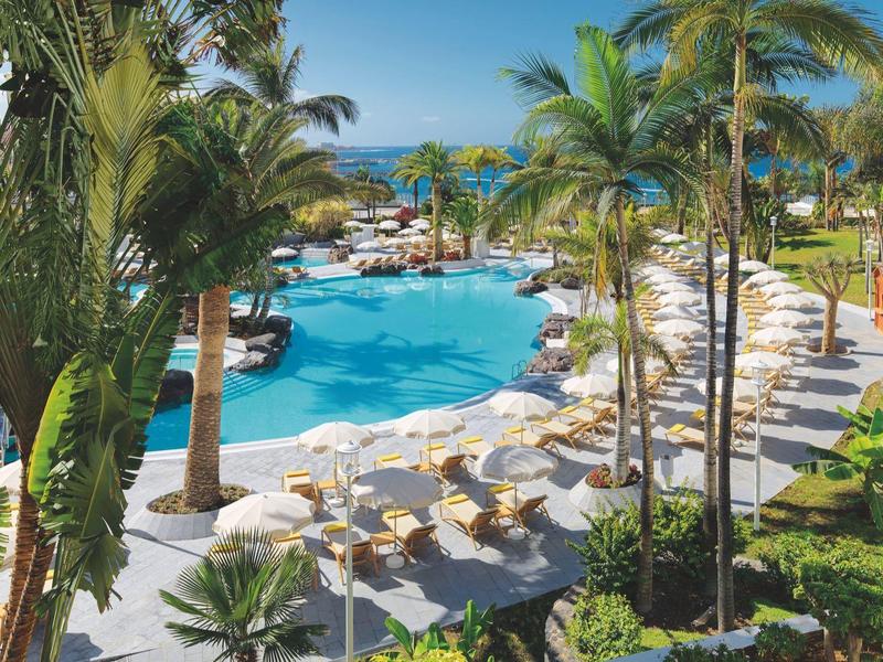 Großer Pool mit Sonnenschirmen, Liegestühlen und Palmen, umgeben von tropischem Garten.