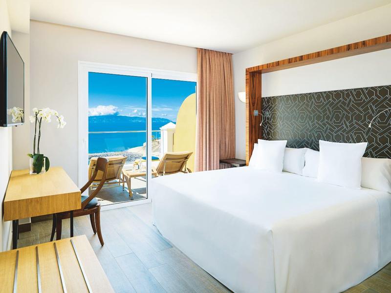 Helles Schlafzimmer mit großem Bett, Holzdekor, Schreibtisch, Pflanzen und Meerblick durch Glasschiebetür.