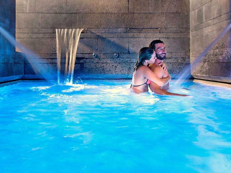 Mann und Frau umarmen sich in blau beleuchtetem Innenpool mit Wasserfall an der Wand.