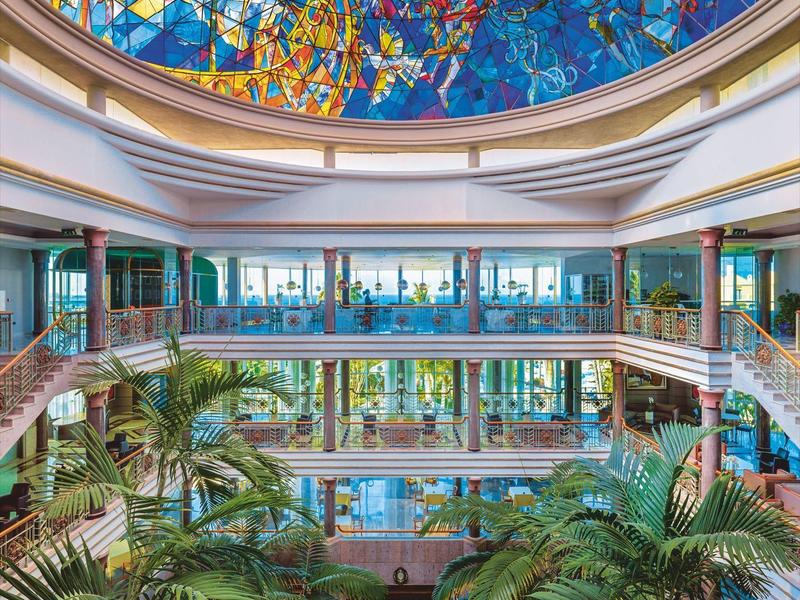 Großes Atrium mit Palmen, Glasgeländer und farbenprächtigem Mosaik an der Kuppeldecke.