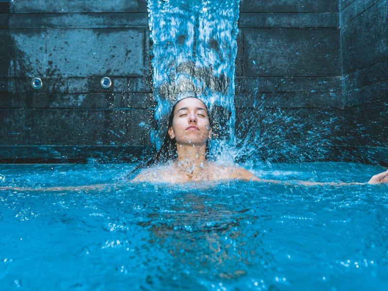 Mann steht unter Wasserfall in klarem blauen Pool, entspannt, mit geschlossenen Augen und leichtem Lächeln.
