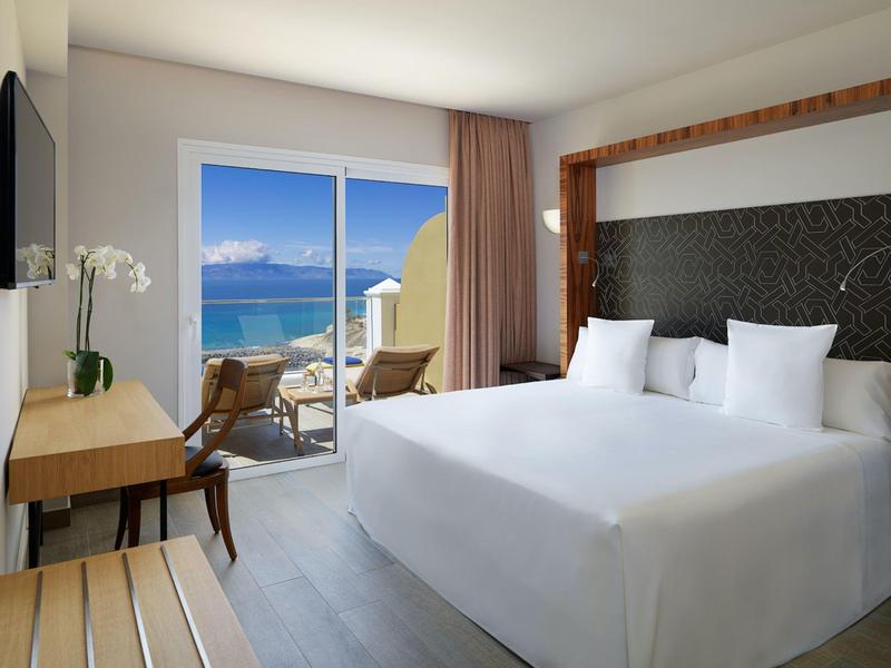 Modernes Hotelzimmer mit großem Bett, Terrasse und Blick auf das blaue Meer bei klarem Himmel.