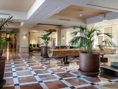 Ruime hotellobby met fluwelen banken, houten tafel en potplanten op een patroonvloer.