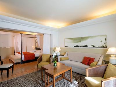 Salon d'hôtel confortable avec canapé, fauteuil, table en bois et lit à baldaquin en arrière-plan.