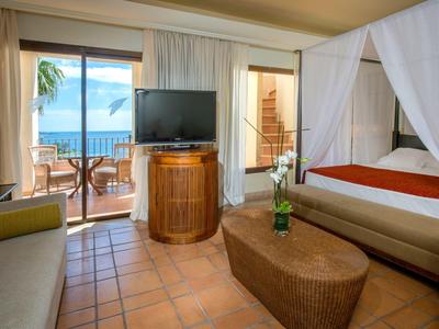 Chambre d'hôtel confortable avec lit à baldaquin, balcon vue océan, TV et mobilier en osier.