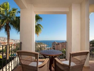 Balcon avec une table ronde, deux chaises, des palmiers et vue sur la mer sous un ciel dégagé.