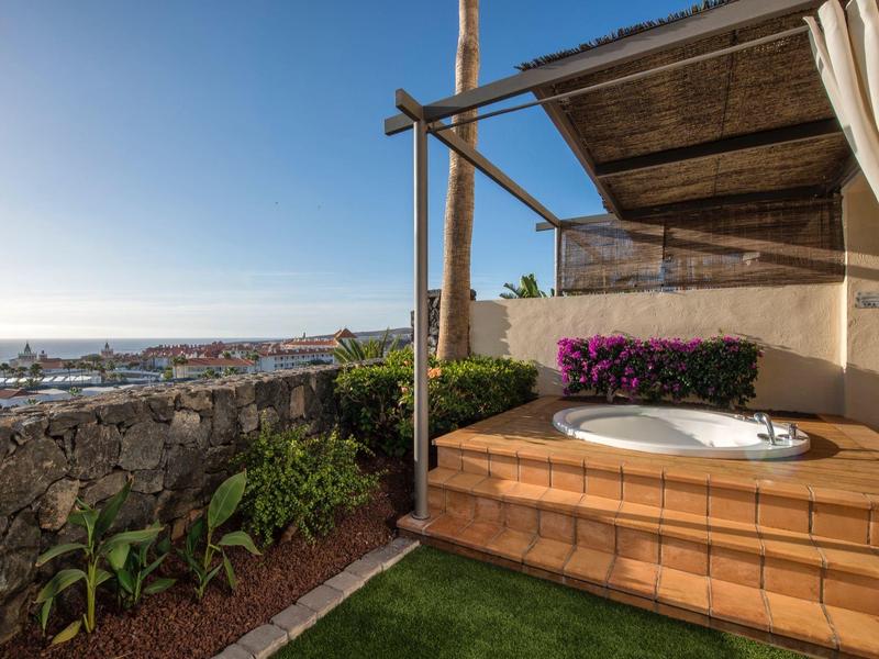 Jacuzzi extérieur sur terrasse carrelée avec vue sur jardin et océan sous un ciel clair.