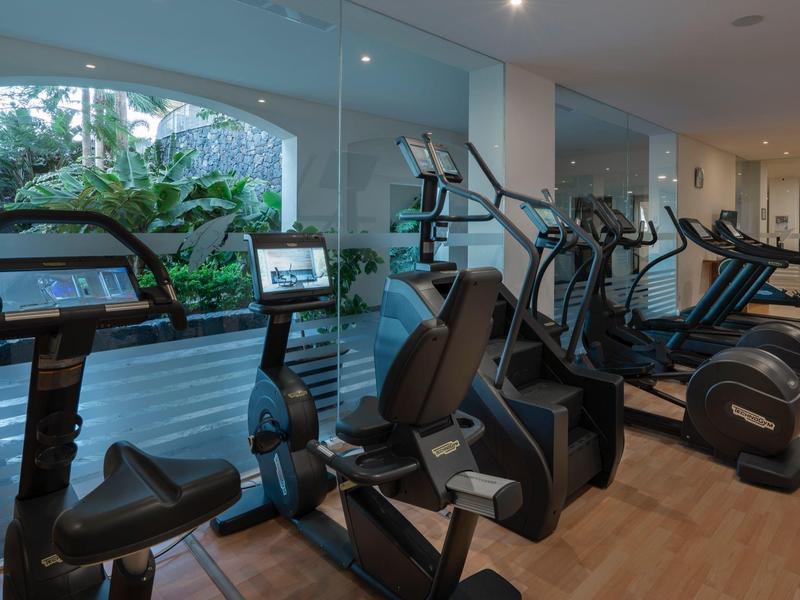 Salle de sport moderne avec vélos d'exercice, tapis de course et grandes fenêtres donnant sur la verdure.