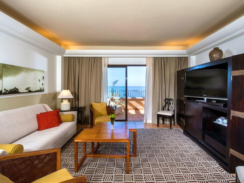 Chambre d'hôtel spacieuse avec canapé, fauteuils, meubles en bois et balcon avec vue sur la mer.