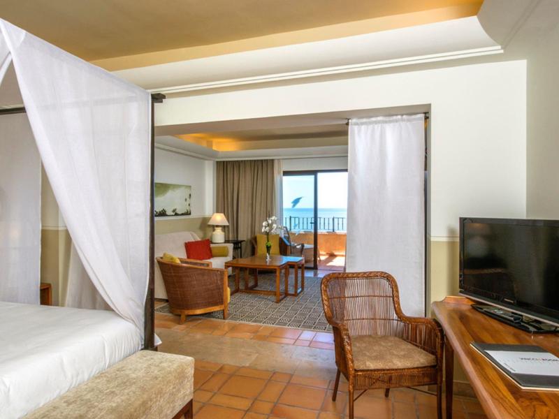 Chambre d'hôtel lumineuse avec lit à baldaquin, chaises en osier, bureau et balcon vue mer.