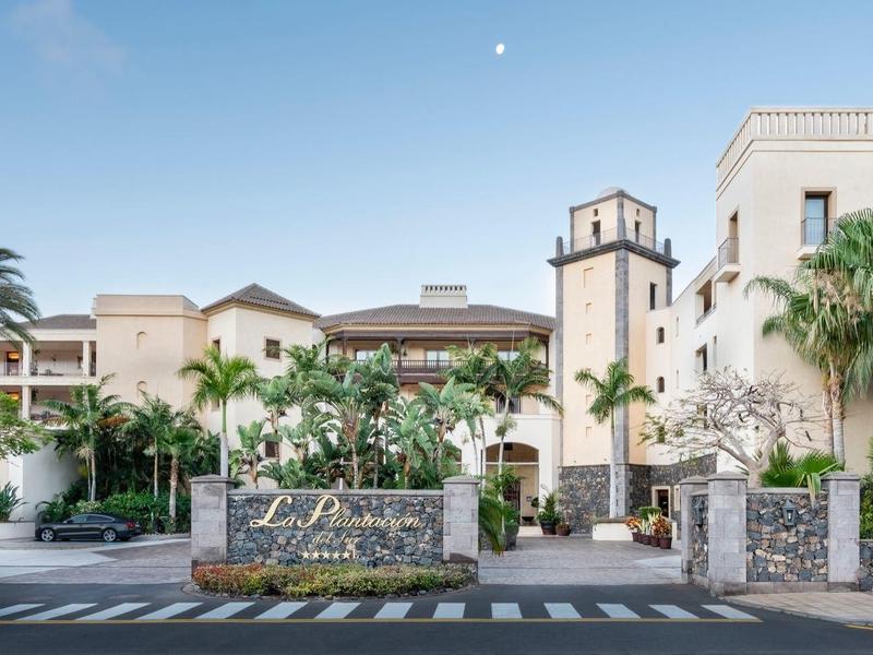 Entrée d'un hôtel de luxe entourée de palmiers et de plantes tropicales sous un ciel clair.