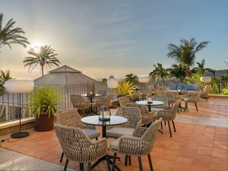Terrasse extérieure d'hôtel avec chaises en osier, tables, plantes et palmiers au coucher du soleil.
