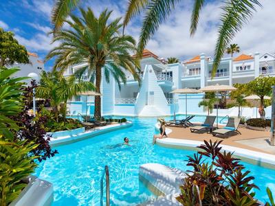 Heller Pool mit Wasserrutsche, Palmen und Liegestühlen vor hellblauen Hotelgebäuden.