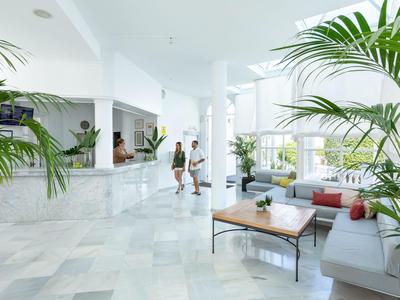 Jasna lobby hotelowa z marmurową podłogą, roślinami i częścią wypoczynkową z stołem i sofami.