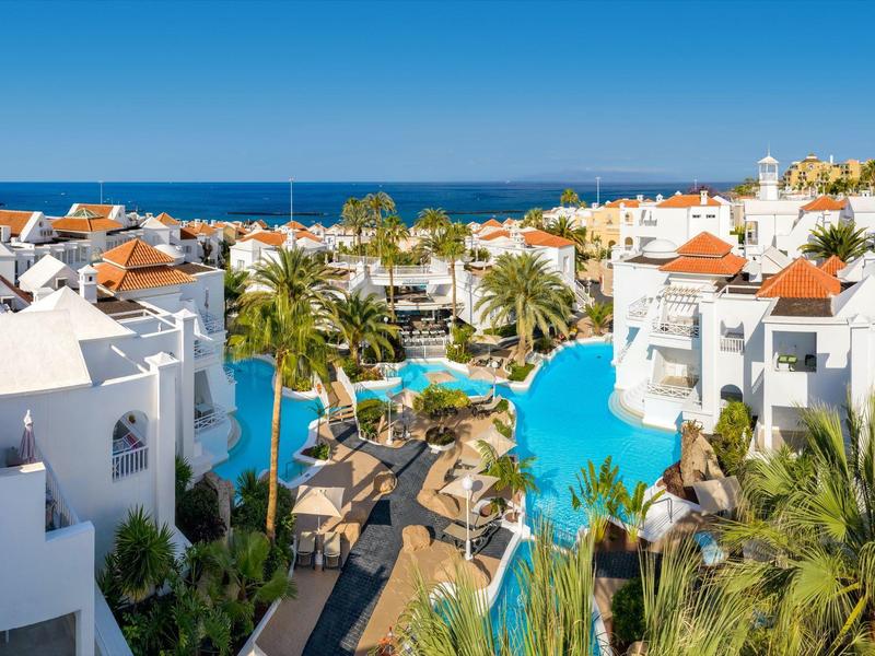 Hotel con edifici bianchi e grande piscina blu, circondato da palme e vista sul mare.
