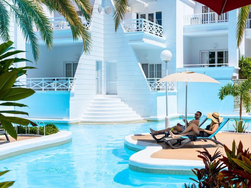 Resort di lusso con piscina, lettini prendisole e piante tropicali sotto il sole.