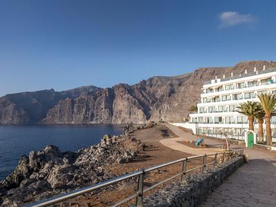 Budynek hotelowy obok skalistego wybrzeża z widokiem na spokojne morze i góry pod jasnym niebem.