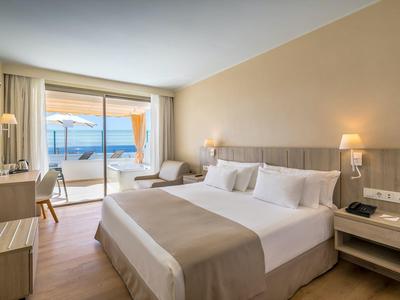 Světý hotelový pokoj s manželskou postelí, posezením a výhledem na moře přes velké skleněné dveře.