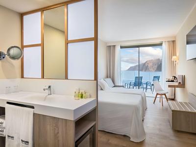 Moderní hotelový pokoj se světlým nábytkem, manželskou postelí, balkonem a koupelnou s velkým umyvadlem.