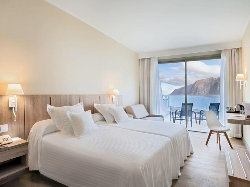Světly hotelový pokoj se dvěma postelemi, posezením a velkým oknem s výhledem na hory.