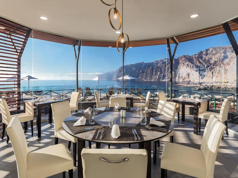 Elegantní restaurace se skleněnými stěnami a pohledem na moře, světlé židle a kulaté dřevěné stoly.