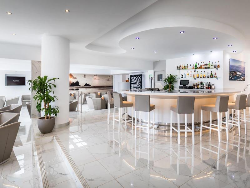 Moderní hotelový bar s bílými židlemi, barovými stoličkami a světlou mramorovou podlahou.