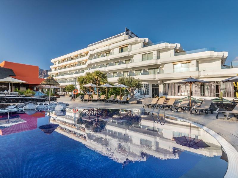 Moderní vícepodlažní hotel obklopený bazénem a lehátky pod jasnou modrou oblohou.