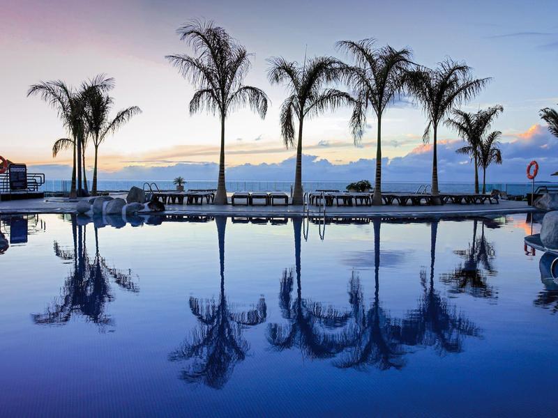 Palmy se odrážejí v klidné vodě bazénu při západu slunce u moře.