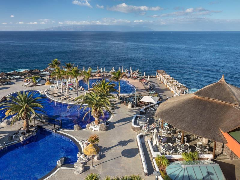 Pohled na hotelový bazén s palmami a mořem v pozadí pod modrou oblohou.