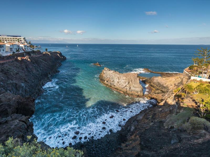 Malá skalnatá zátoka s vlnami, obklopená budovami a vegetací u moře.