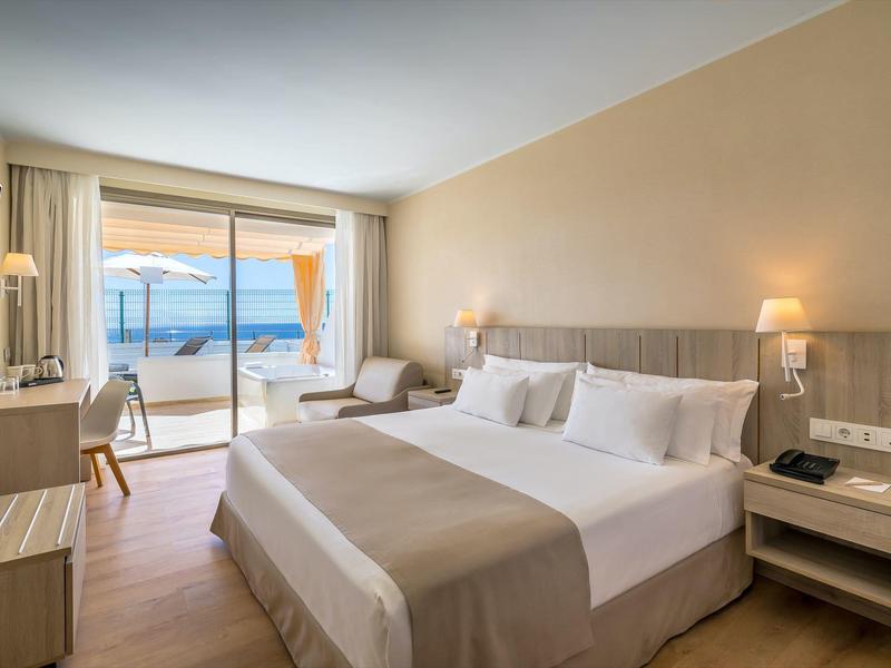 Světý hotelový pokoj s manželskou postelí, posezením a výhledem na moře přes velké skleněné dveře.