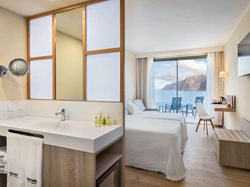 Moderní hotelový pokoj se světlým nábytkem, manželskou postelí, balkonem a koupelnou s velkým umyvadlem.