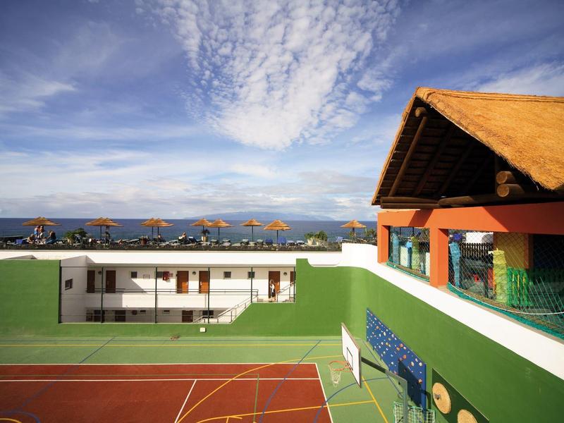 Pohled na tenisové kurty a budovu s terasou v hotelovém komplexu s horami.