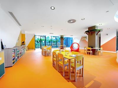 Heldere kinderspeelkamer met oranje vloer, kleine tafels, stoelen en speelelementen.