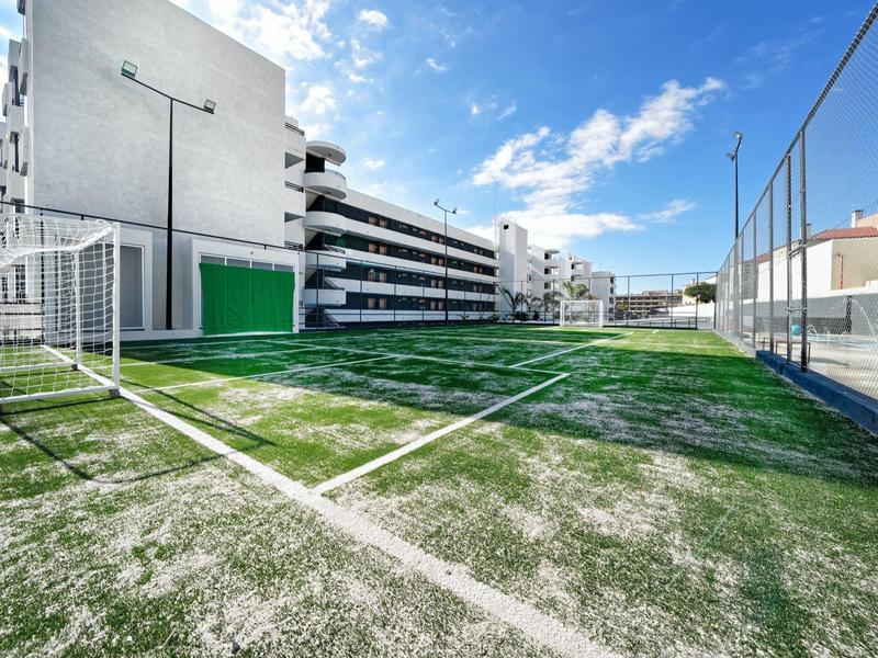 Klein sportveld met kunstgras naast een wit meerlaagse gebouw onder een blauwe lucht.