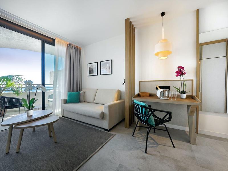 Modern hotelkamer met bank, tafel, bureau en balkon met zeezicht.