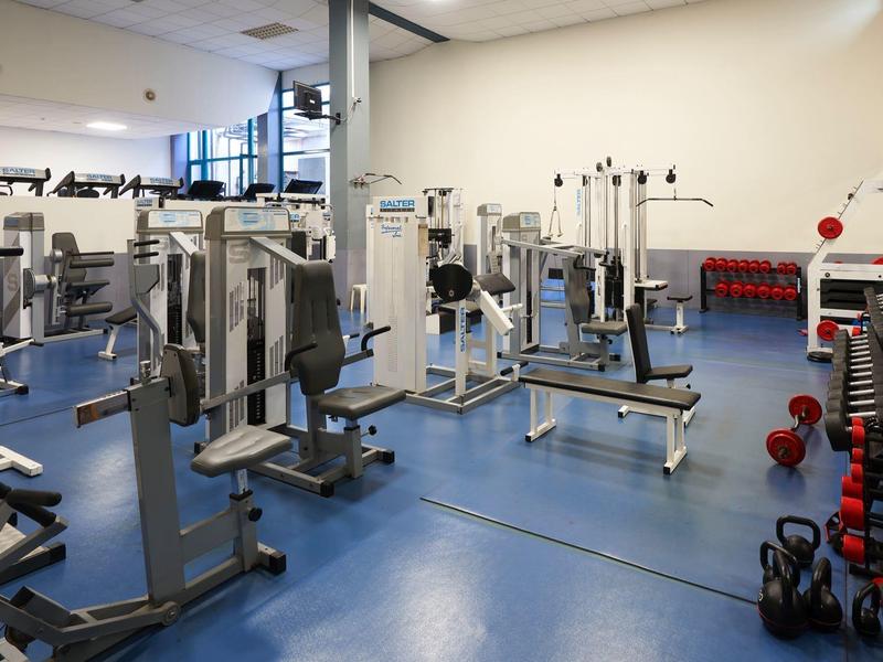 Ruime sportschool met diverse fitnessapparaten en gewichten op een blauwe vloer.