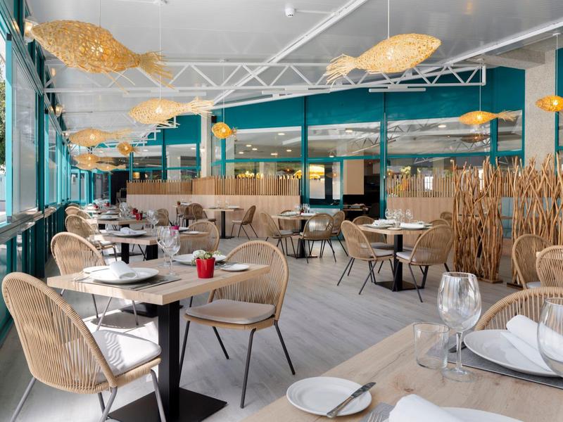 Lichte hotel eetzaal met rotan stoelen, ronde tafels en grote ramen met uitzicht op groen buiten.