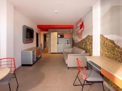 Moderně zařízený hotelový pokoj s pohovkou, psacím stolem, židlemi a kuchyňským koutem.