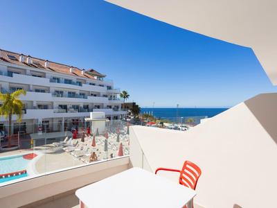 Balkon s stolem a červenými židlemi s výhledem na hotelový bazén a moře za jasné oblohy