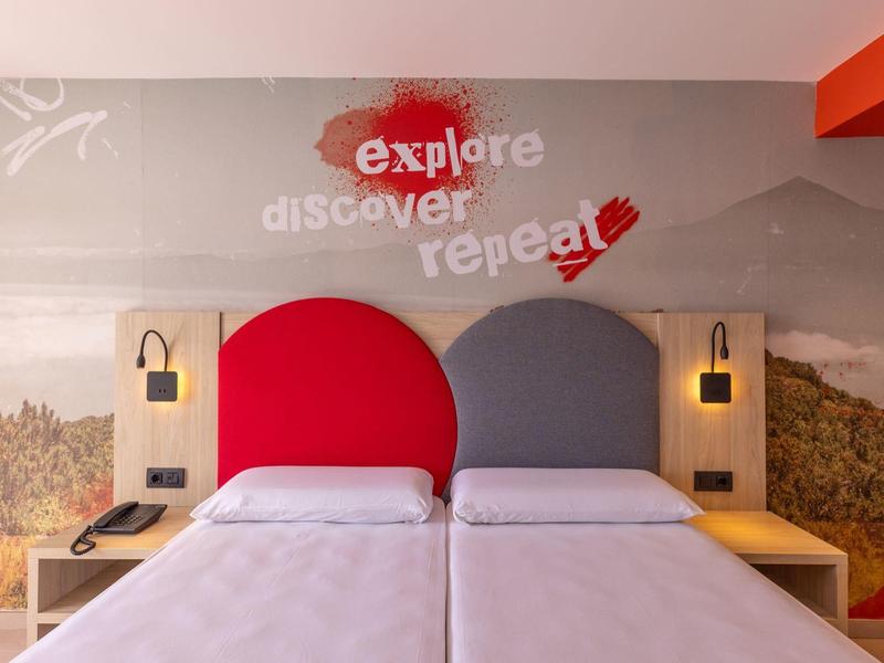 Hotelový pokoj se dvěma postelemi, červeným a šedým čelem postele, moderní nástěnnou dekorací a čtecími lampami.