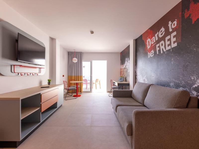 Moderní hotelový pokoj s pohovkou, TV, psacím stolem a balkonem se denním světlem.