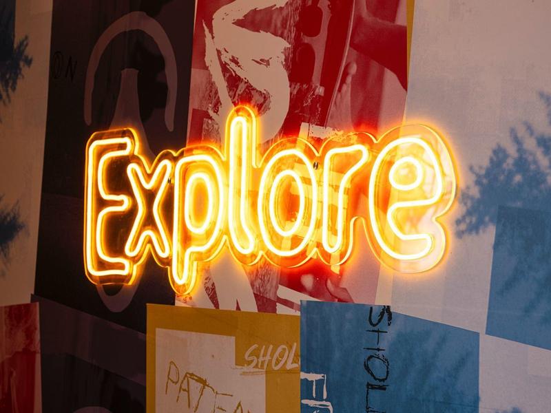 Neonový nápis 'Explore' na barevné vzorované zdi s stíny.