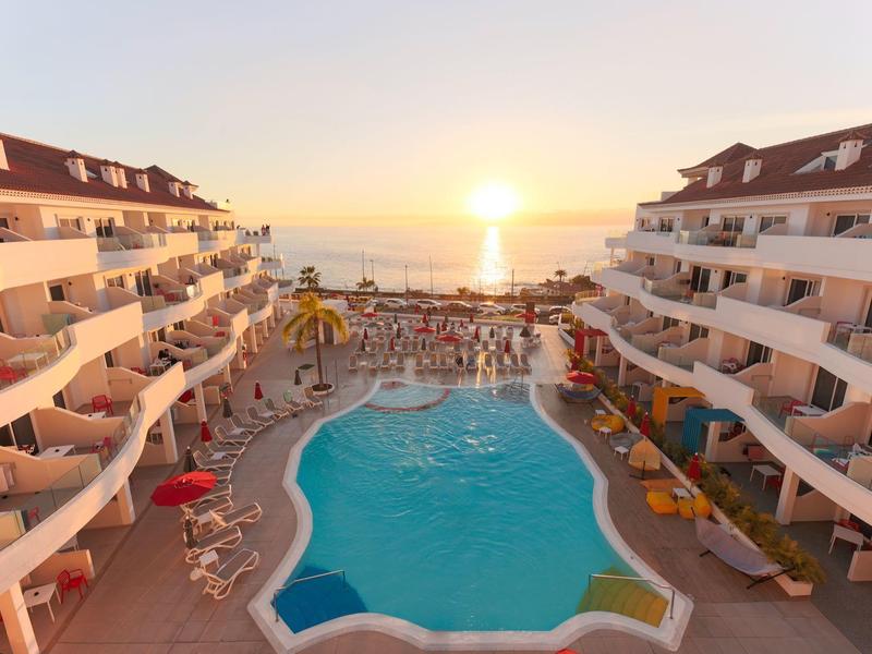 Pohled na hotelový bazén mezi dvěma budovami při západu slunce s výhledem na moře.