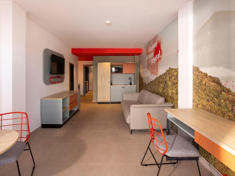 Moderně zařízený hotelový pokoj s pohovkou, psacím stolem, židlemi a kuchyňským koutem.