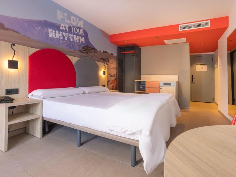 Moderní hotelový pokoj s manželskou postelí, červeným čelem postele a barevným designem stěn.